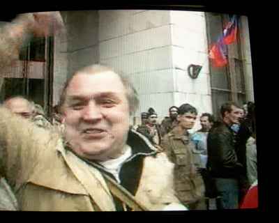 one man, face near camera, waving one arm in the air, looking happy with a small crowd of men behind him paying no attention
